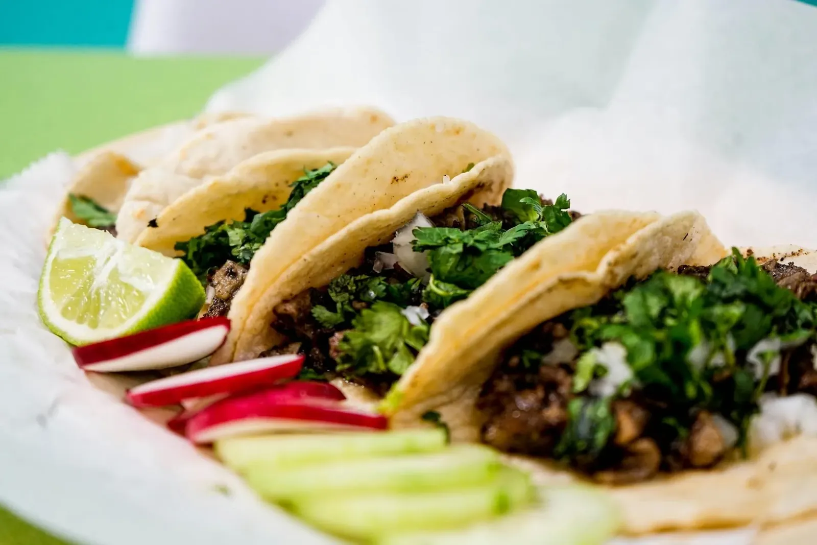 Three tacos with cilantro and onion in a paper-lined basket, with a lime wedge and sliced radish