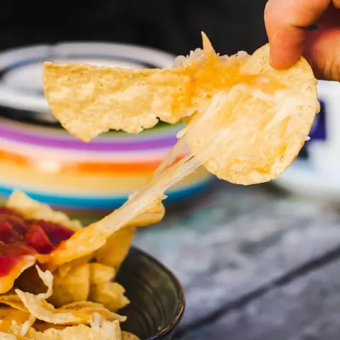 A hand pulling a stretchy melted-cheese tortilla chip from a bowl of nachos with red salsa