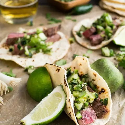 Steak tacos topped with chopped onion and cilantro, with lime halves on parchment paper