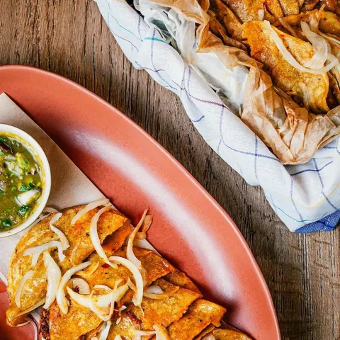 Rolled, fried tortillas topped with onions on an oval terracotta plate, beside a small dish of green salsa