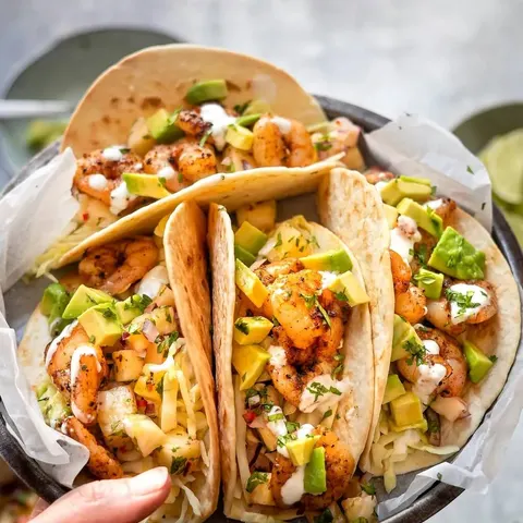 A hand holding a plate of three tacos filled with grilled meat, avocado slices and a creamy drizzle