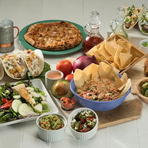 A spread of tacos, salad, chips, guacamole, salsa and two pizzas on a pale wood surface